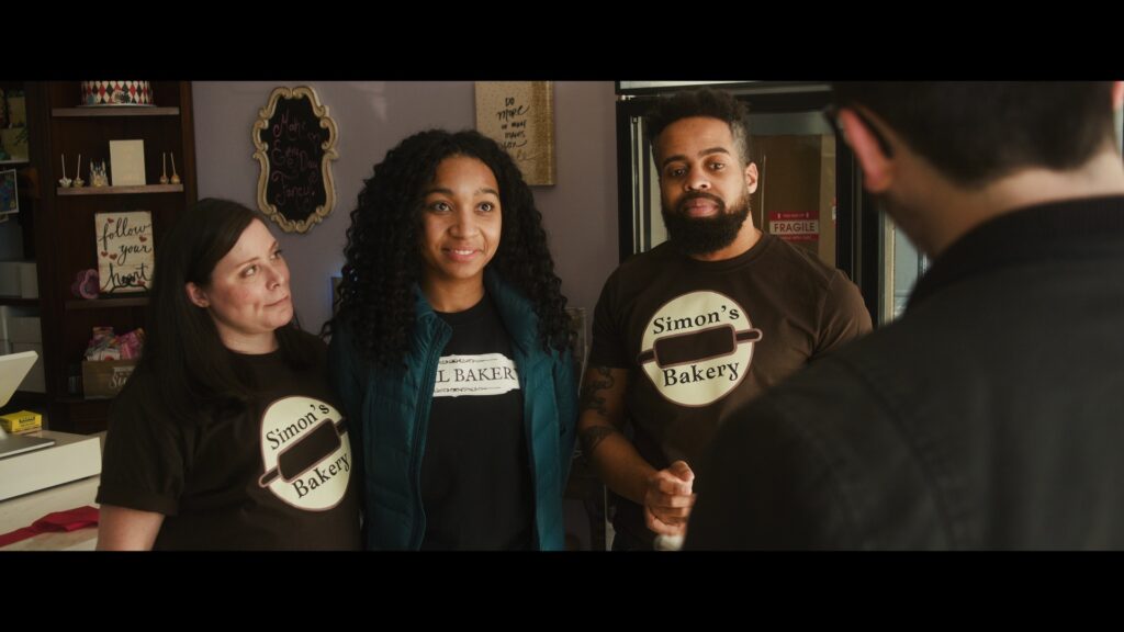 A still image from Annie's film showing a group of three people wearing shirts for "Simon's Baker"  talking to another person who is seen from behind from the shoulder up.