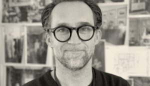 Faculty member Curt Beech stares into the camera in an artful black and white photo with out of focus pictures hanging on a wall behind him
