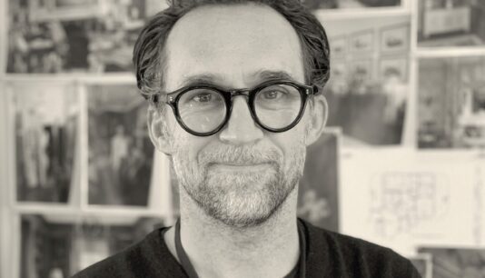 Faculty member Curt Beech stares into the camera in an artful black and white photo with out of focus pictures hanging on a wall behind him