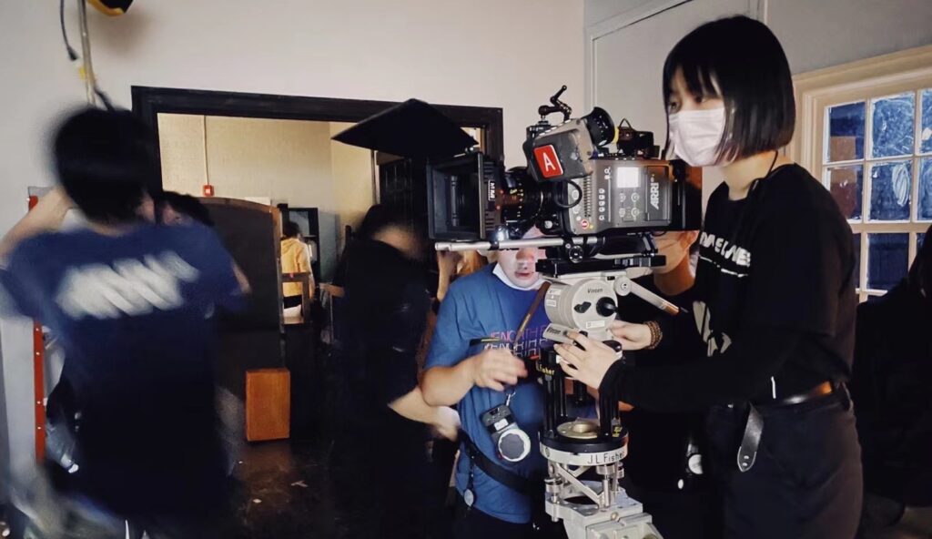 behind the scenes photo shows a group of crew members on set working. one person with a mask operated a camera while others work around them.