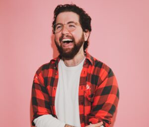 Oliver Slate Green stands in front of a solid pink backdrop wearing a checkered red and black flannel over a white shirt smiling at the camera.