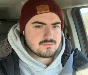 Student Chris Stager takes a selfie in a car wearing a red beanie and a hoodie under a jacket.