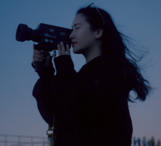 Director Lamu Xiangqiu stands in profile holding a camera up to her eye against the backdrop of the dark blue night sky.