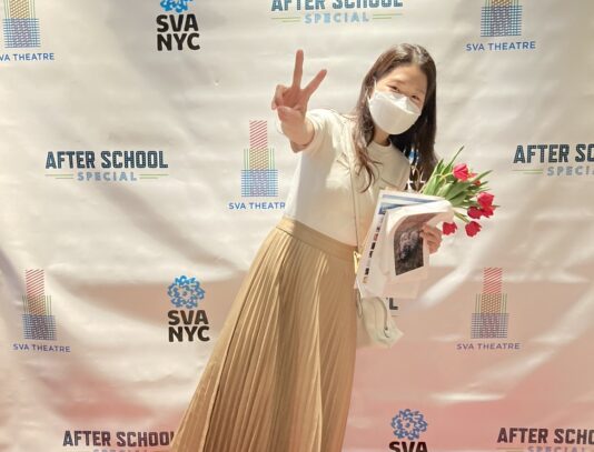 Julie Jang stands in front of the SVA After SChool Special Red carpet with a bouquet and hodling their hand up in a peace sign.