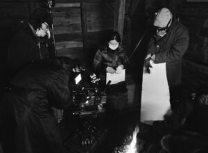 Inside of a log cabin, four people stand near the camera and prepare for the next take.