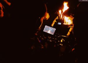 A crew member holds the camera very close to a fire pit.