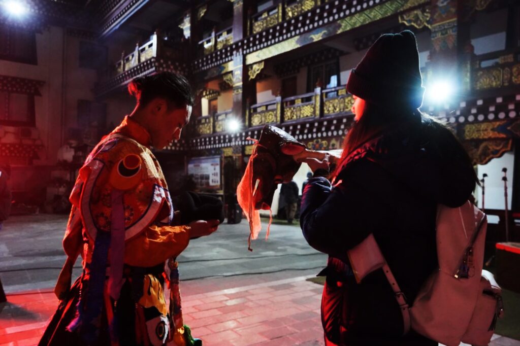 behind the scenes photo of an actor looking down at their phone while a crew member offers them an animal mask.