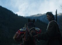 A person with a weapon on their back is feeding a horse in front of a mountain.