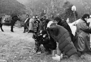 The cast and crew are outside, setting up the camera for a shot. Three crew members hover behind the camera, while an actor with a horse is seen in the background.