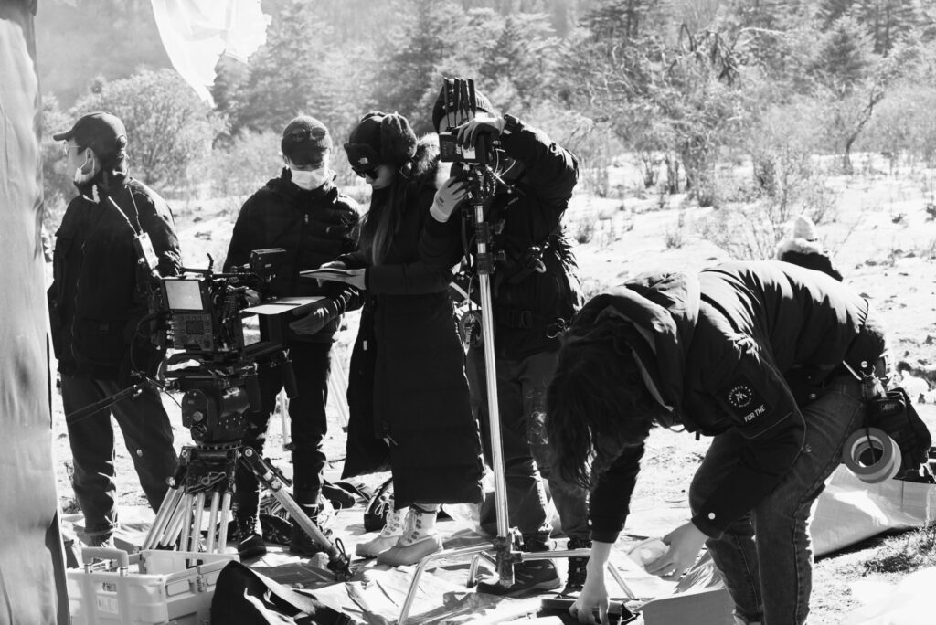 Five crew members who are outdoors are preparing their equipment to shoot a scene. A large camera on a tripod and a c-stand are with them.