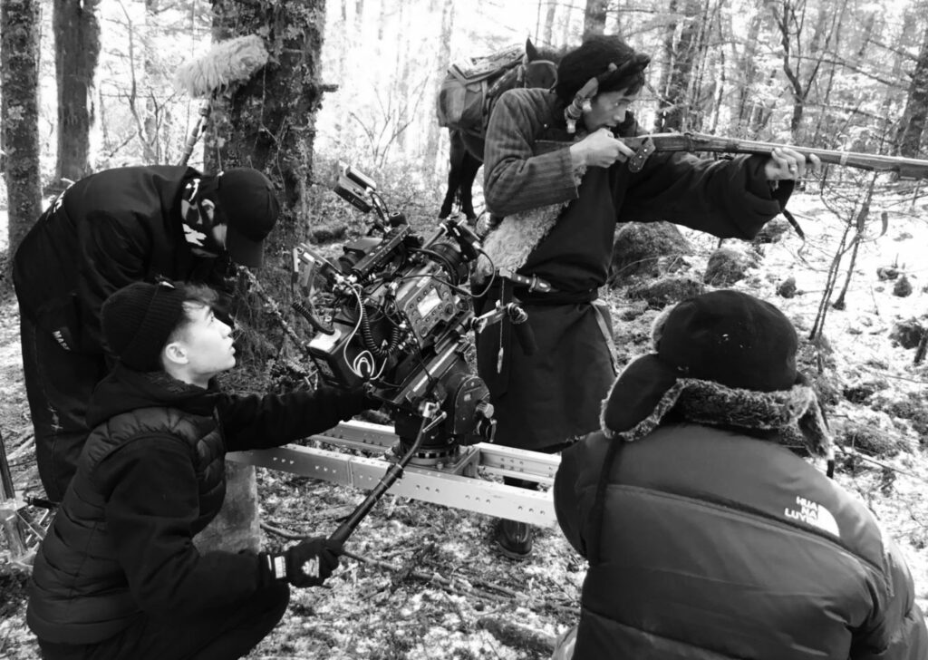 A crew member is pointing the camera to an actor while the actor is holding a weapon. Two other crew members stand next to the camera operator.