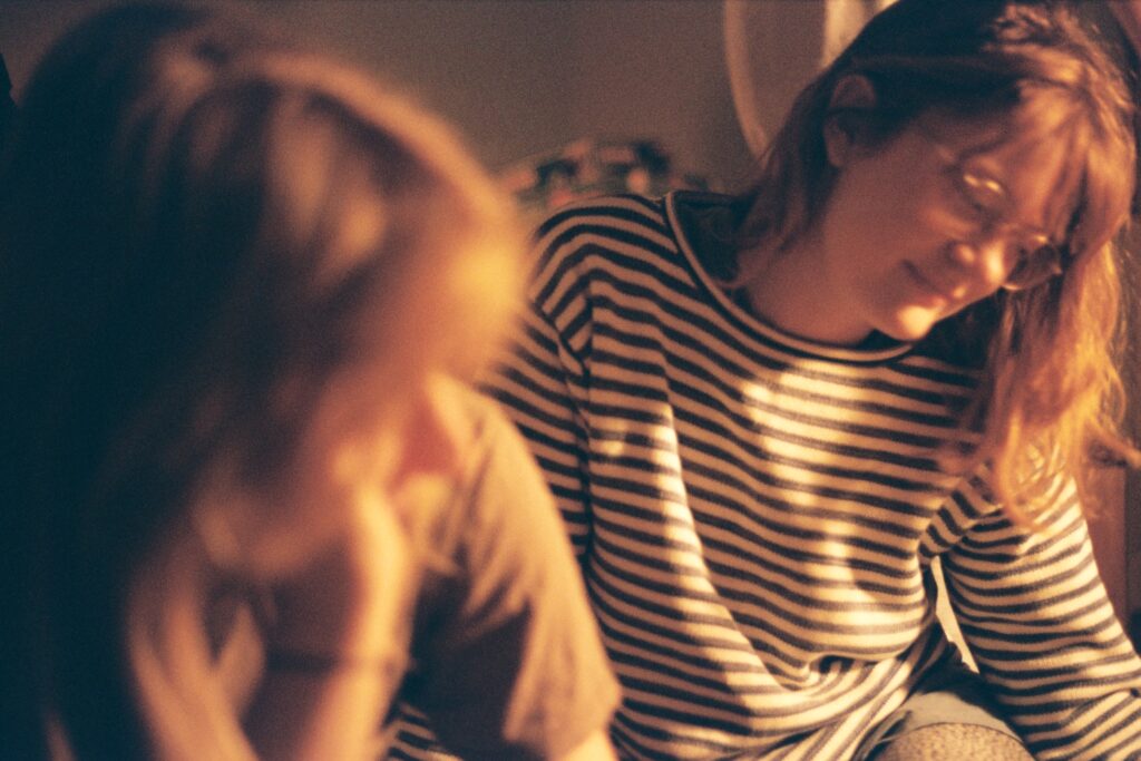 a person in a striped shirt and glasses looks down toward the ground and another person out of focus is turned back to look at them.