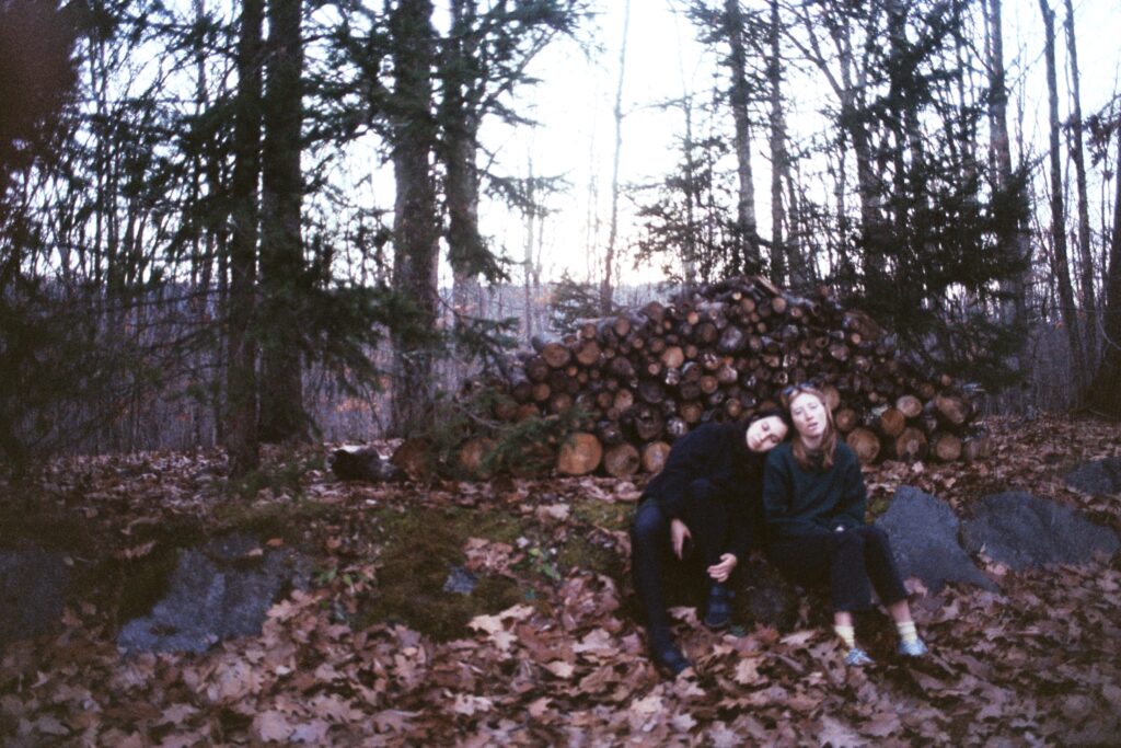 Gonnie Zur and another person sit outside in a forest on a pile of leaves with barren trees behind them..