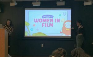 Two students from the Women in Film Club stand in front of a projector with the words "Welcome to: Women in Film"