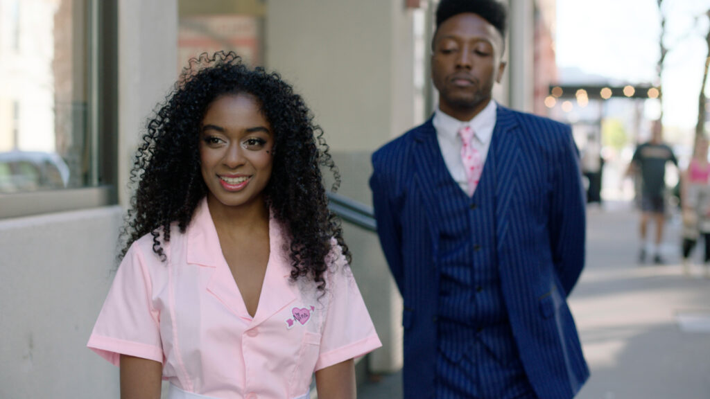 A person in a pink jumpsuit stands next to a well dressed person in a blue suit and pink tie.