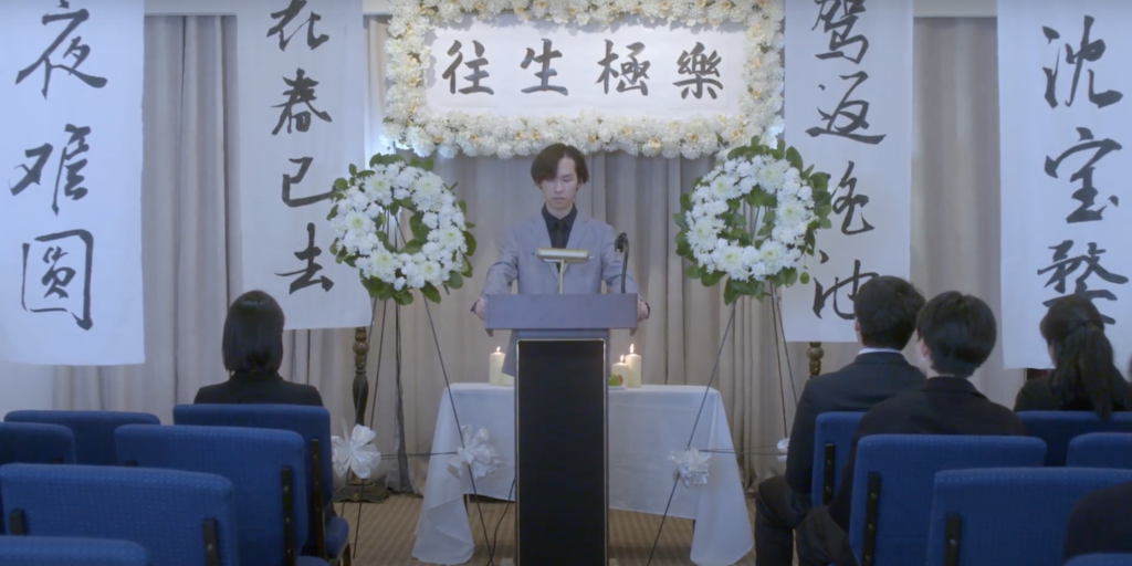 a person speaks at the podium at a funeral home while attendees sit and watch