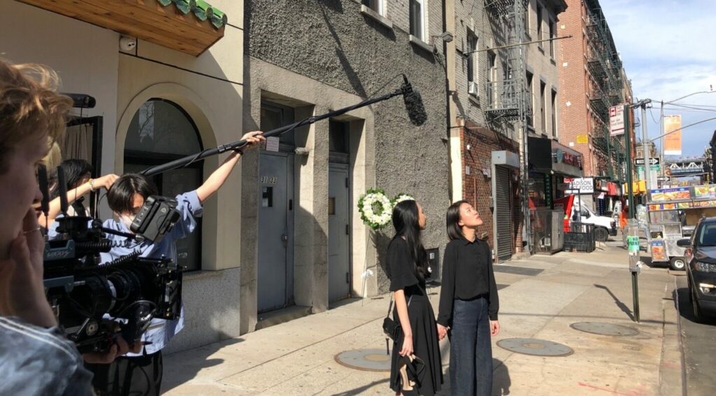 a behind the scenes photo from the set outside the funeral home. Shows the two actors looking off camera while crew members photograph and capture sound off to the side.
