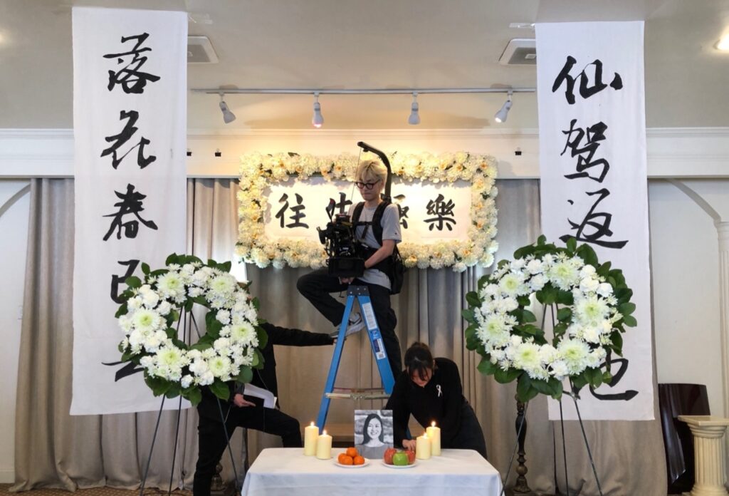 A behind the scenes photo from the set of the funeral home. Film crewmembers prepare a shot with a cameraperson in the middle of the photo on a ladder and floral arrangements surround them and two other people setting up the props.