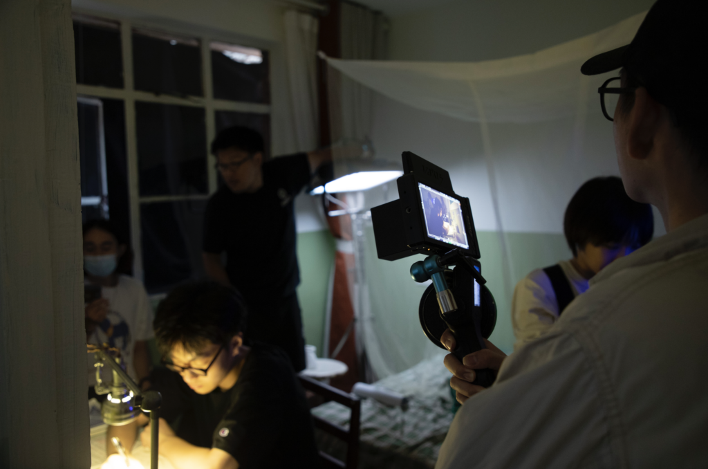 behind the scenes photo from Tulpa showing several crew members setting up a shot of an actor at a desk. Another crew member looks through the viewfinder of the camera.
