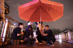 behind the scenes photo from Tulpa showing several crew memnbers setting up a shot outside under an umbrella. Most are bent down gathered around a camera.