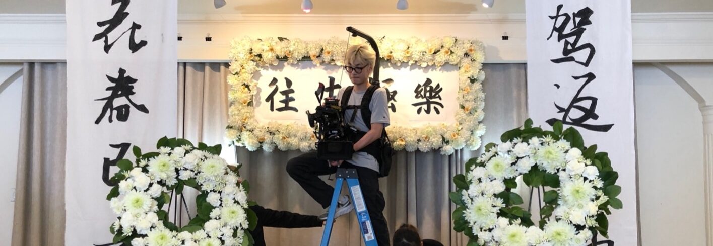 A behind the scenes photo from the set of the funeral home. Film crewmembers prepare a shot with a cameraperson in the middle of the photo on a ladder and floral arrangements surround them and two other people setting up the props.