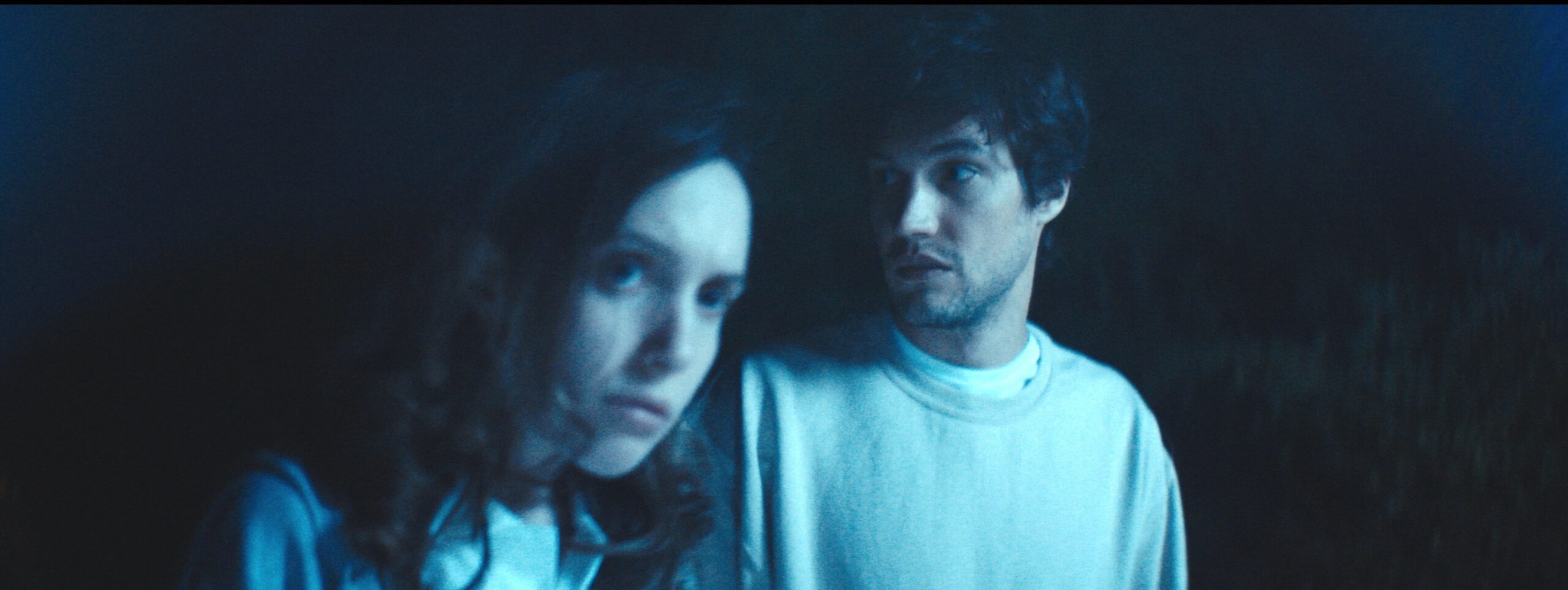 A still from Robert McMahon's film showing two people in close up bathed in a blue light with black bacgkround.