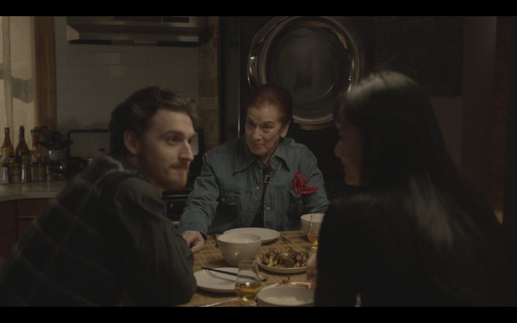 A trio of people sit at a dining room table with food on the table in a dimly lit apartment.