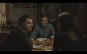 A trio of people sit at a dining room table with food on the table in a dimly lit apartment.