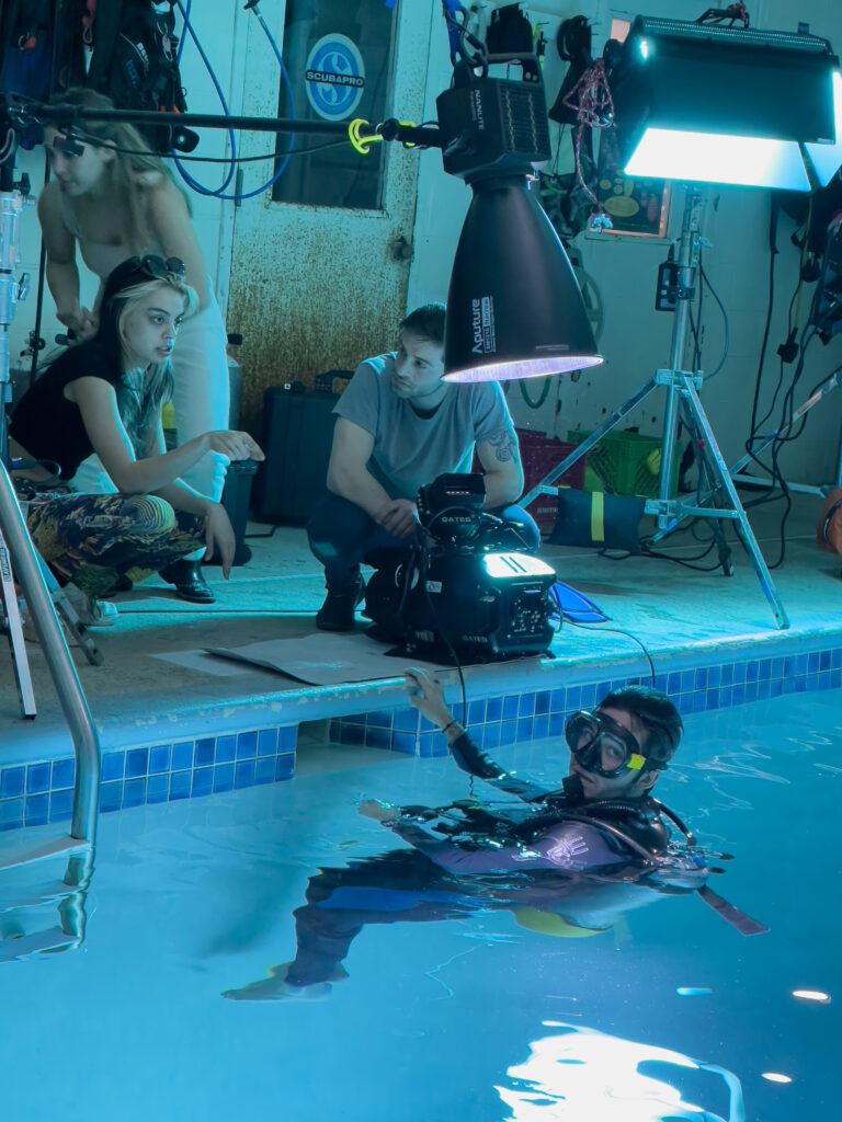 Crewmembers on Lainne's film prepare for an underwater scene with several crewmembers gathered around the edge of a pool while another in full diving gear floats in the water.