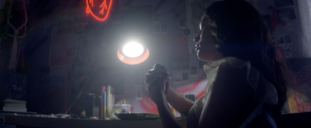 Still from Lianne's film showing a young wmoan sitting at her desk in her room with a light from a desklamp shining on her.