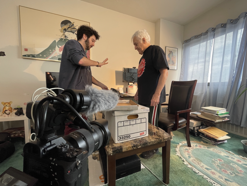 Kenny and Ray behind the scenes of filming with camera equipment set up for an interview. Both standing.