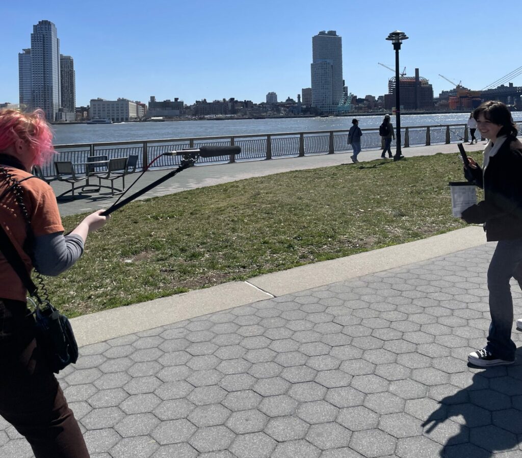 Slate about to be clapped on set on a street by the water while another crew member holds out a microphone on a boom pole.