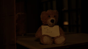 The same teddy bear from the second picture, now propped up alone on the same furniture. You can see a bit of his reflection in the mirror behind him. There is a piece of paper with loopy handwriting on his belly, that reads "you'll never be alone.