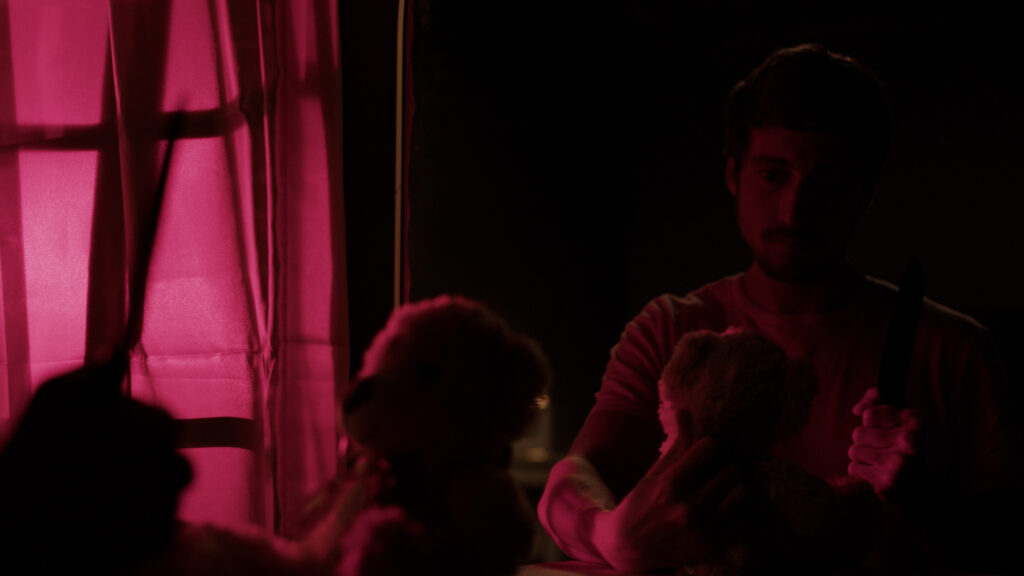 In a dark room, an actor holds a teddy bear in one hand and a knife in the other. He looks at it aggressively. There's a window shining red light onto his face and a mirror in front of him, the photo is taken from the reflection of it.