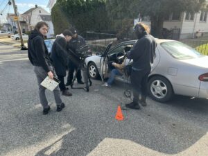 Behind the scenes photo from Louis's film showing the crew standing outside of a car they are preparing to shoot a scene with.