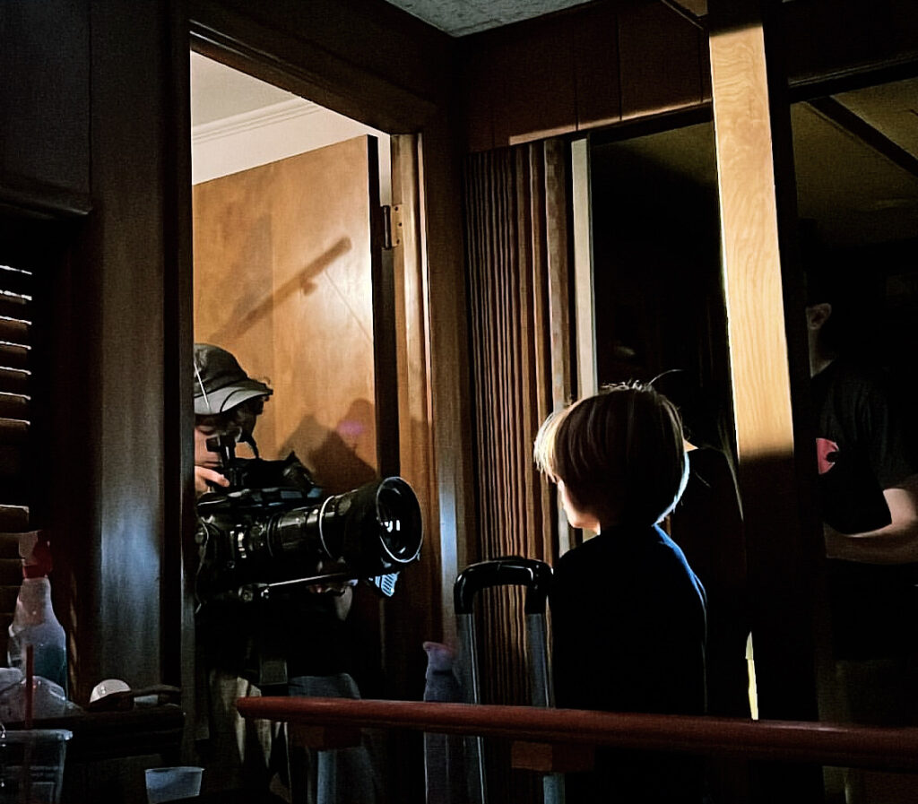 a child actor standing in a dark room, a cinematographer holding a camera in front of the child actor