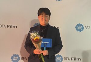 Alumnus Louis Yeo stands in front of an SVA BFA Film Step and Repeat holding flowers smiling