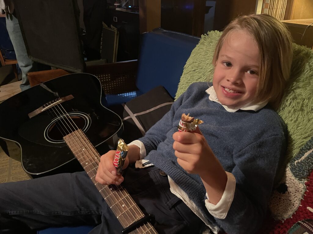 A child actor siting on a chair with a guitar eating chocolate snack
