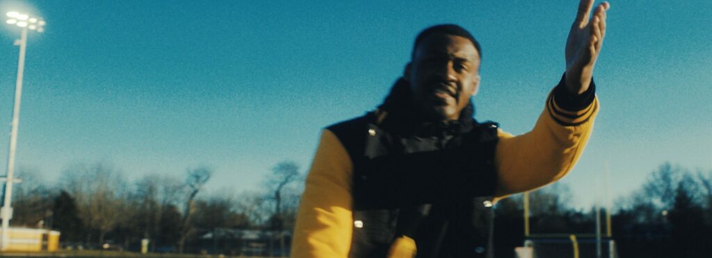 Screenshot from Khalil's film Pass Da Mic shows an actor on a football field smiling and raising their hand.