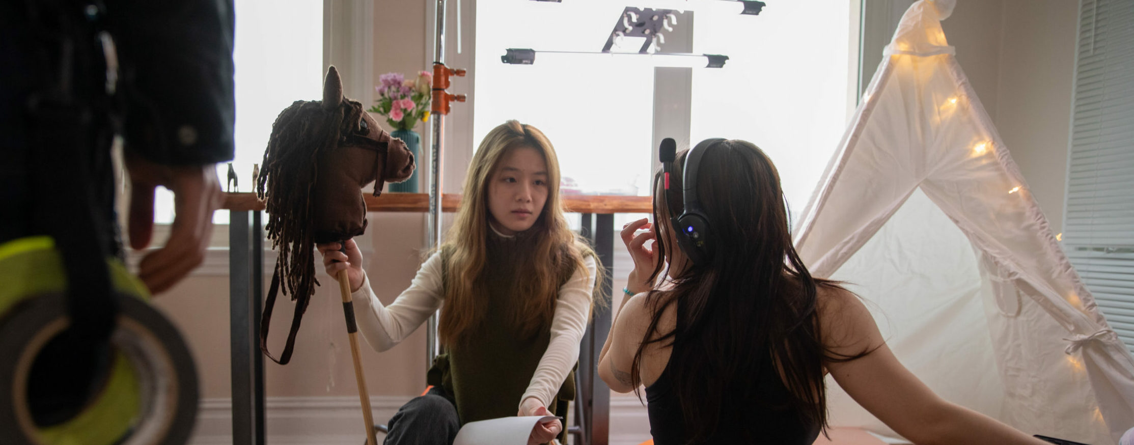 Mina crouches on set holding a hobby horse head and pole while talking with a fellow crewmember.