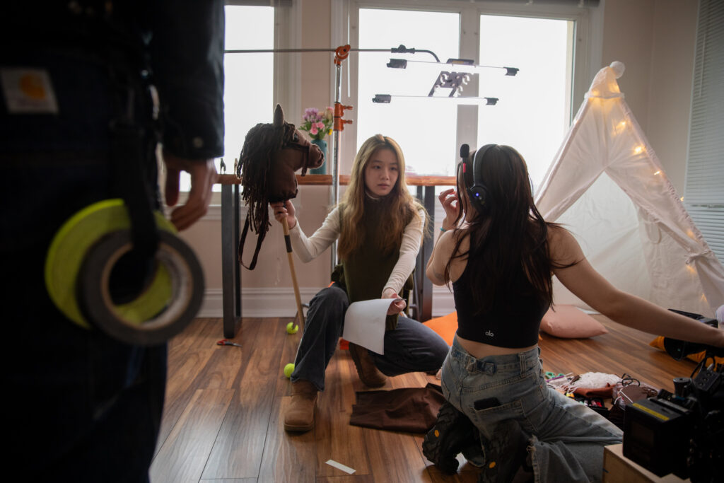 Mina crouches on set holding a hobby horse head and pole while talking with a fellow crewmember.