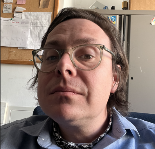 Headshot of a man in an office wearing a neckerchief and blue shirt and glasses.