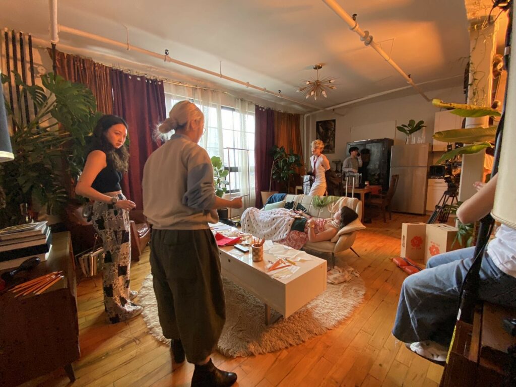 Filmmakers in a cluttered and colorful living room, an active film set. There is film equipment around, and they seem to discuss what the next steps in production are.