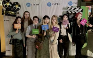 The crew of Lucky Shot is in front of an SVA BFA Film banner, dressed up. They smile to the camera and hold plaques that display their work on set. Director Zixia (Jessica) Cheng is in the middle.