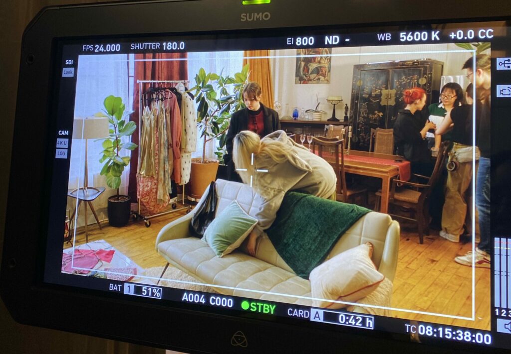 Image of a film monitor recording a scene from Zixia's film showing actors and crew preparing for a scene in an apartment with a couch and dining room table and big curtains.