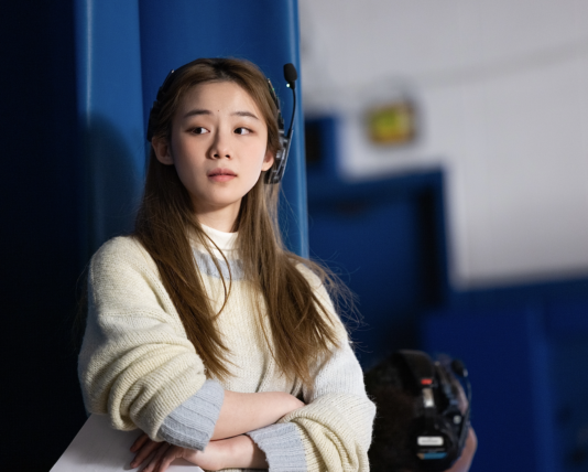 Director Mina Huang stands on set with arms crossed wearing a headset.