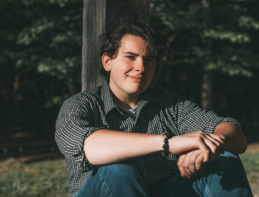 Young man sitting on a wooden porch, leaning his hands over his knees and his back against a wooden column.