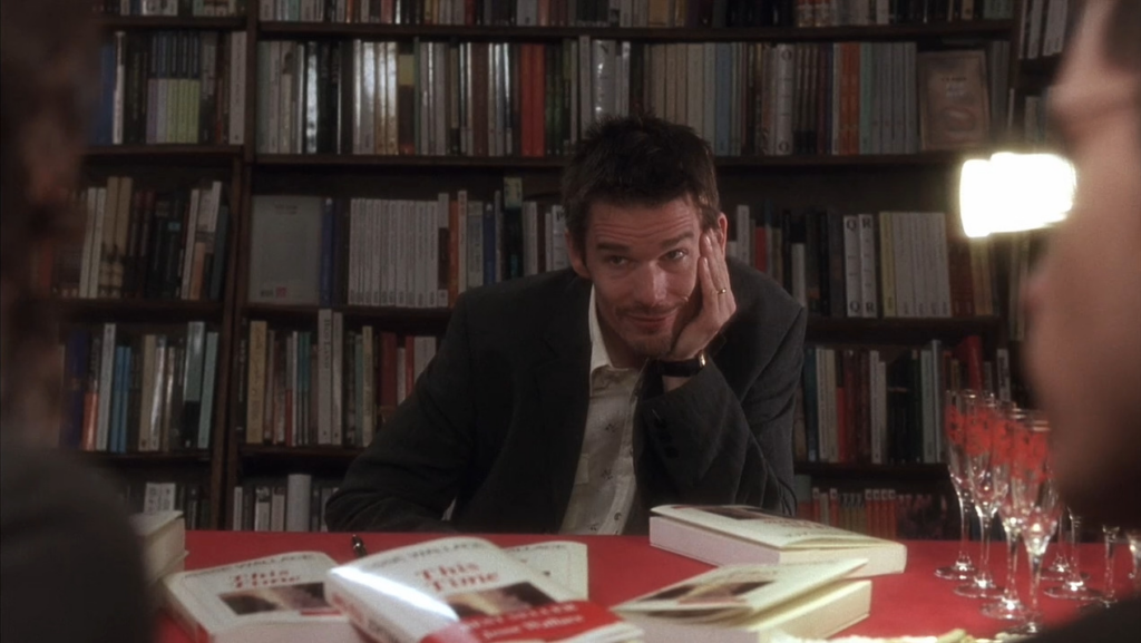  A young man wearing a suit sits behind a red table with books. He looks to a couple talking to each other, who are barely in frame. Behind him, there are shelves of books.