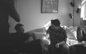 Black and white film photo. Director Matt Haller sits on a couch with actor Simon Henriques as the crew works around them. They discuss the next scene.