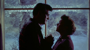  A couple holds hands and looks into each other's hands. Behind them is a big glass window lighting them from behind, casting dark shadows over their faces.